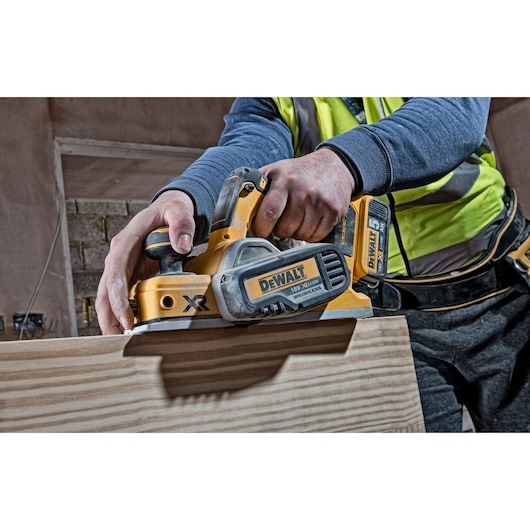 A person using a DEWALT DCP580N cordless planer to smooth the surface of a wooden board. The tool is yellow and black, with visible branding and battery.