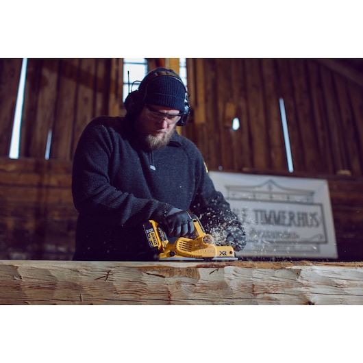 A person using a yellow DEWALT cordless planer (model DCP580P2) to smooth a large piece of wood inside a workshop. The person is wearing safety earmuffs, gloves, and a dark sweater.