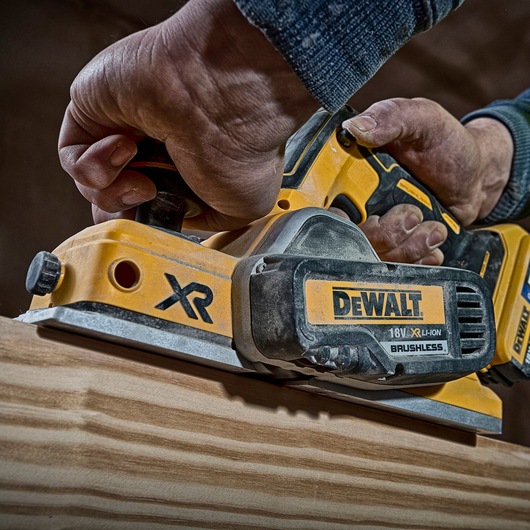 Hand uses a DEWALT DCP580P2 cordless planer to smooth wood. Close-up of tool and wood surface.