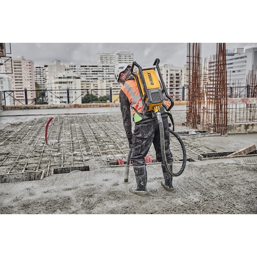 A construction worker wearing a safety helmet and vest operates a DEWALT backpack concrete vibrator on a building site