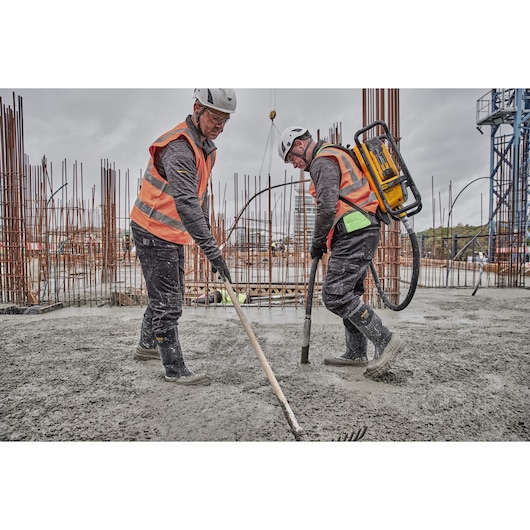 Two construction workers wearing safety helmets and orange reflective vests are working on a concrete surface. One worker is