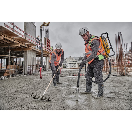 Two construction workers wearing safety helmets and vests are working on a concrete floor at a construction site. One worker