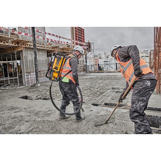 Two construction workers wearing safety helmets and orange vests working on a concrete floor at a building site. One worker