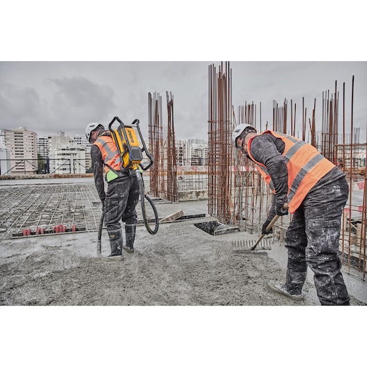 Two construction workers wearing safety helmets and orange vests are working on a concrete slab at a construction site. One