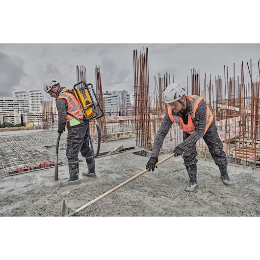 Two construction workers wearing safety helmets and orange vests are working on a concrete slab at a building site. One