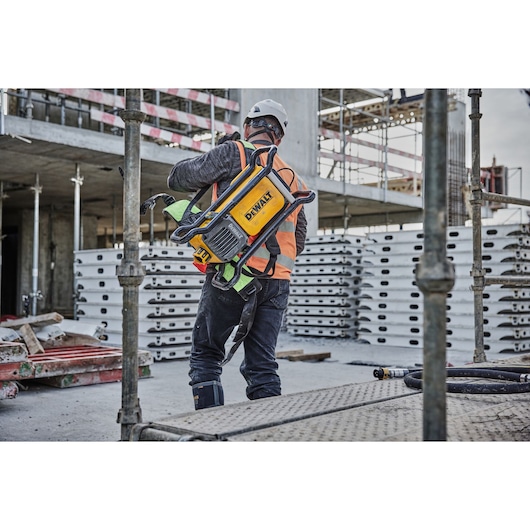 A construction worker wearing safety gear and a hard hat is carrying a DEWALT DCPS310N power tool on their back at a construction site.