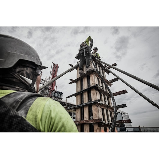 Construction workers wearing safety gear are working on a tall wooden structure at a job site. One worker is seen from the