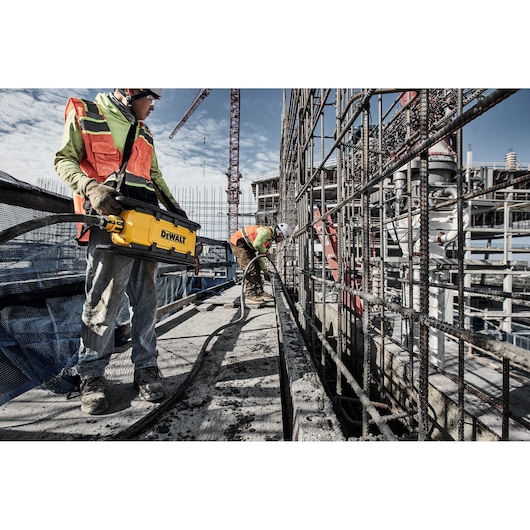 Two construction workers are wearing safety vests and helmets at a building site. One worker is holding a yellow DEWALT