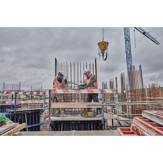 Two construction workers wearing safety helmets and orange vests are working with steel rebar on a building site