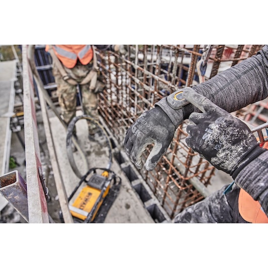 Worker wearing gloves operates DEWALT tool at a construction site, surrounded by metal rods and safety equipment.