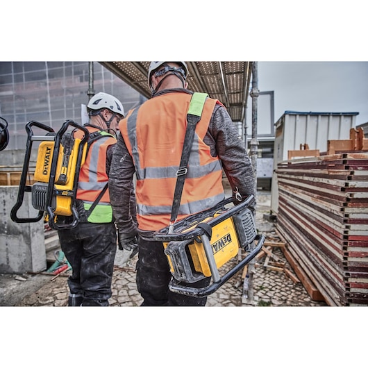 Two workers in safety gear carry DEWALT equipment at a construction site with scaffolding and materials.
