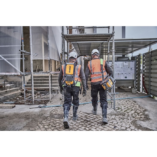 Two workers with DEWALT equipment walk at a construction site wearing helmets, boots, and orange safety vests.