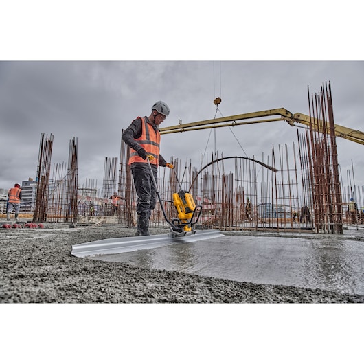 A worker wearing safety gear and a high-visibility vest uses a DEWALT DCPS330N power tool to smooth wet concrete on a construction site. Surrounding the worker are steel reinforcement bars and other construction equipment, with cloudy skies overhead.