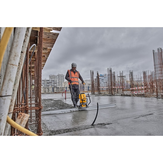 A construction worker wearing a safety vest and helmet uses a DEWALT power tool to smooth wet concrete on a construction site. Metal rods and urban buildings are visible in the background under a cloudy sky.