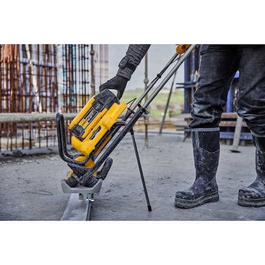 A DEWALT cordless pole saw being operated on a construction site, with a worker wearing gloves and protective boots standing nearby. The tool is yellow and black and is being used to cut at ground level. Construction materials and steel bars are visible in the background.