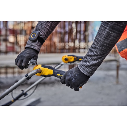 Close-up of a worker adjusting a DEWALT DCPS330N cordless pole saw outdoors, wearing safety gloves and a grey long sleeve shirt with an orange safety vest.