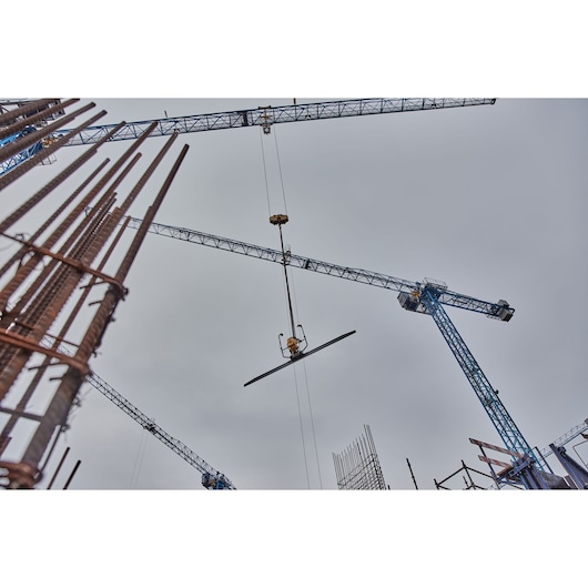 Construction site with several blue tower cranes lifting a metal beam against a cloudy sky, surrounded by steel reinforcement bars and framework.