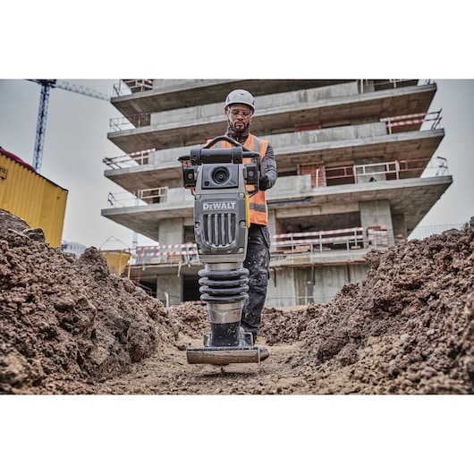 A construction worker wearing a safety helmet and orange vest operates a DEWALT compactor on a dirt pathway at a