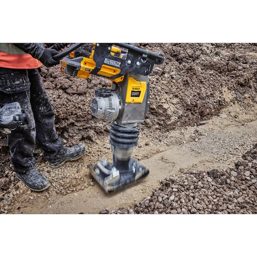 A DEWALT rammer compactor is being used to compress soil on a construction site. The machine is yellow and black, and a