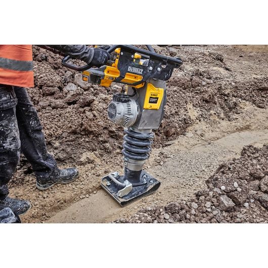 A person operating a DEWALT battery-powered compactor on a construction site, compacting soil with the machine.
