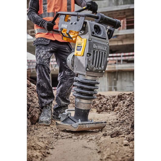A person wearing a high-visibility vest operates a DEWALT rammer compactor on a construction site, compacting soil in a
