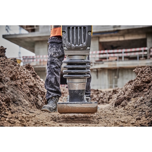 Close-up of a DEWALT compactor tool being used by a worker to compress soil at a construction site, with dirt mounds and