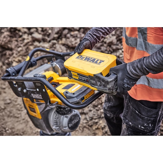 Worker installs DEWALT battery pack onto a yellow DEWALT outdoor power tool at a construction site.