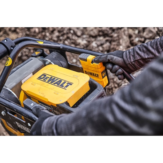 Person with gloves operates a yellow DEWALT power tool outdoors, focusing on controls and battery area.
