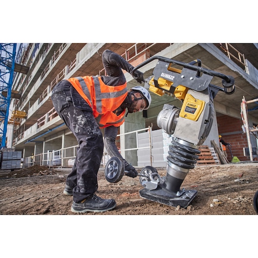 Worker on construction site uses a yellow DEWALT compactor tool while adjusting its wheel attachment.