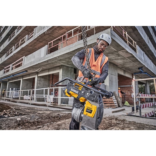 Worker adjusting a DEWALT tool on a construction site, wearing safety gear and helmet near a building structure.