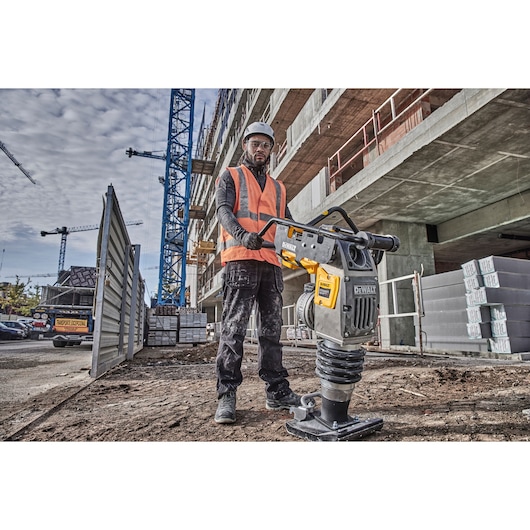 Worker using a DEWALT compactor at a building site, wearing safety gear. Construction materials are visible nearby.