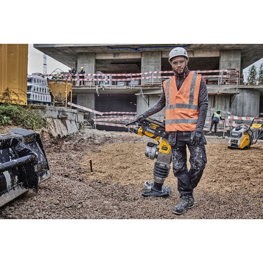 Worker in safety gear using a DEWALT compactor at a construction site with machinery and building in the background.