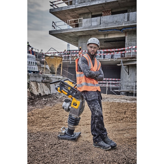 Worker in orange vest and helmet stands next to a DEWALT compactor at a construction site.