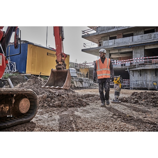 Worker in safety vest and helmet using DEWALT tool at a construction site with excavator and building in background.