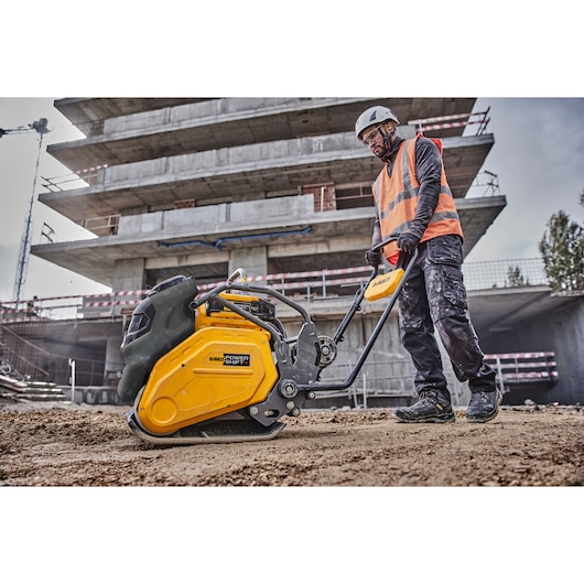 A person wearing a safety helmet and an orange reflective vest operates a yellow DEWALT POWER SHIFT plate compactor on a