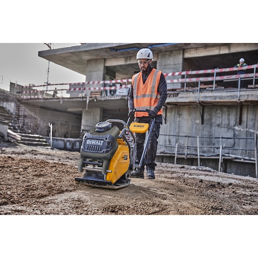 A construction worker wearing a high-visibility orange vest and helmet operates a DEWALT plate compactor on a dirt surface