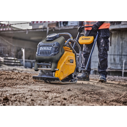 A DEWALT plate compactor being used on a construction site, operated by a person wearing work boots, dark pants, gloves, and
