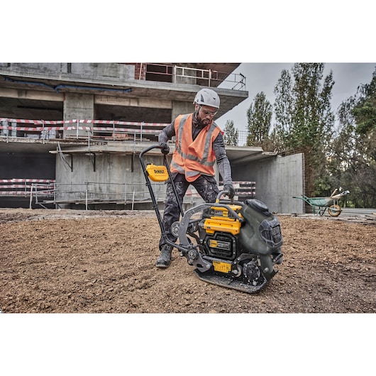 A construction worker wearing a helmet and orange safety vest operates a DEWALT DCPS7154N plate compactor on a dirt surface