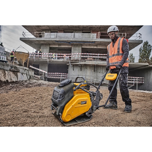 A construction worker operates a DEWALT plate compactor on a dirt surface at a building site, with a partially constructed