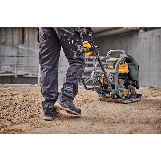 A person operating a DEWALT plate compactor on a construction site, smoothing soil or gravel. The machine is yellow and