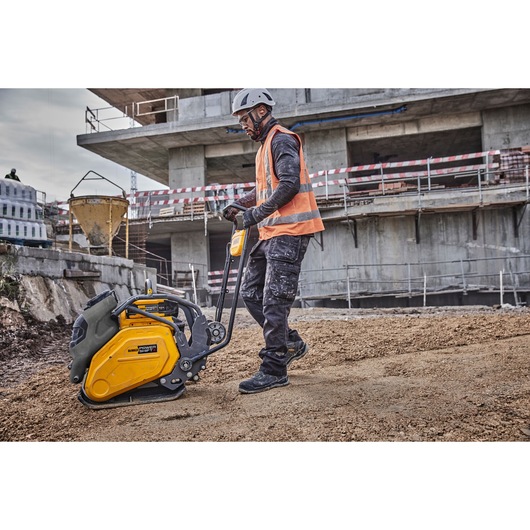 A construction worker wearing a helmet and an orange safety vest operates a yellow DEWALT plate compactor on a dirt surface