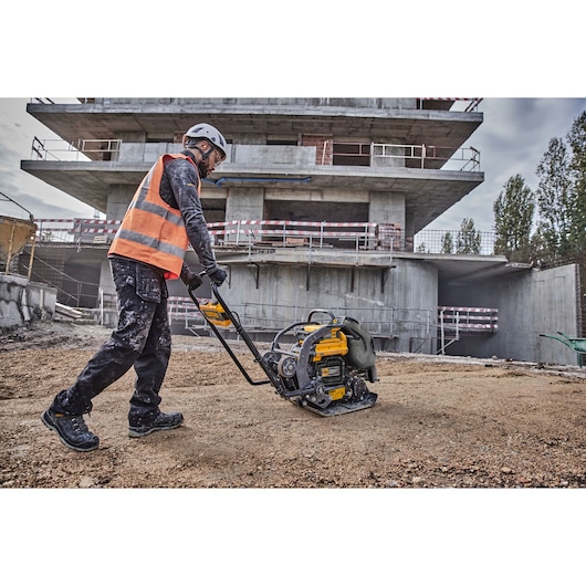 A construction worker wearing a safety helmet, high-visibility vest, gloves, and protective boots is operating a DEWALT