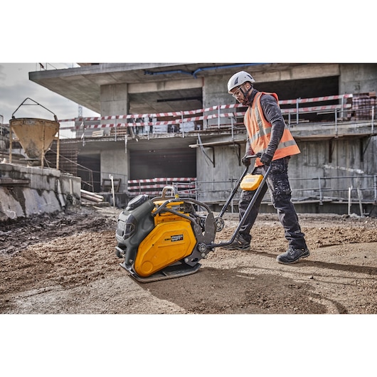 A construction worker wearing a safety helmet and orange vest operates a yellow DEWALT compactor machine on a sandy surface