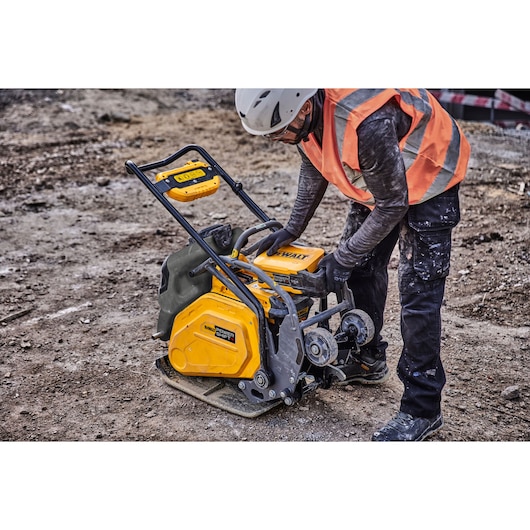 Worker in safety gear operates a yellow DEWALT plate compactor on a construction site.