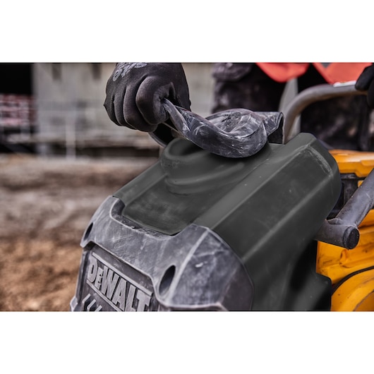 Gloved hand lifting the cover of a DEWALT tool outdoors, showing part of the tool's body and handle.