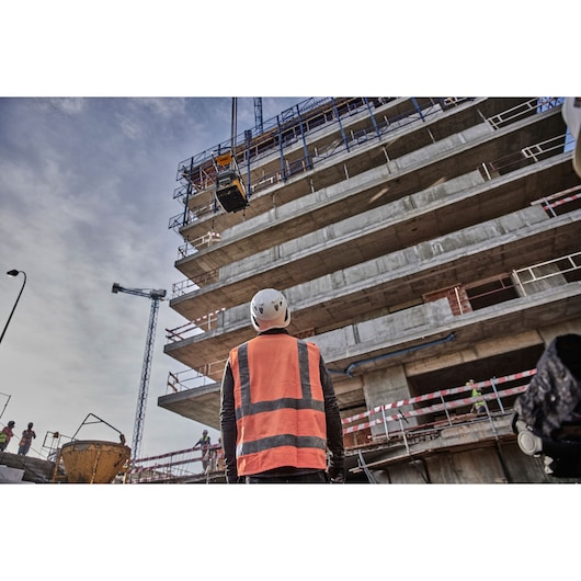 Construction worker in safety gear looks up at building under construction with equipment being lifted by crane.
