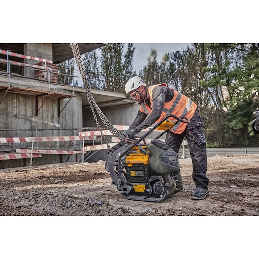 Worker in safety gear handles a DEWALT compactor at a construction site with dirt and chain equipment visible.