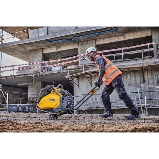 Person uses a DEWALT plate compactor on a construction site, wearing a helmet and high-visibility vest.