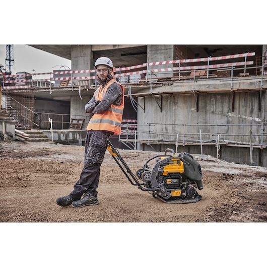Worker in safety gear leans on a DEWALT plate compactor at a construction site with concrete and scaffolding.