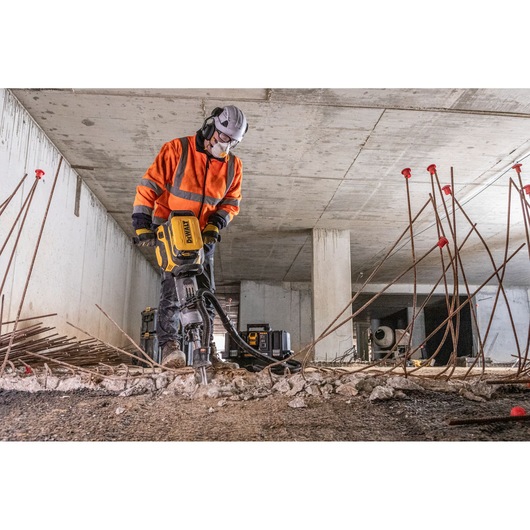 Worker using DEWALT concrete breaker tool indoors with safety gear amid rebar and rubble.